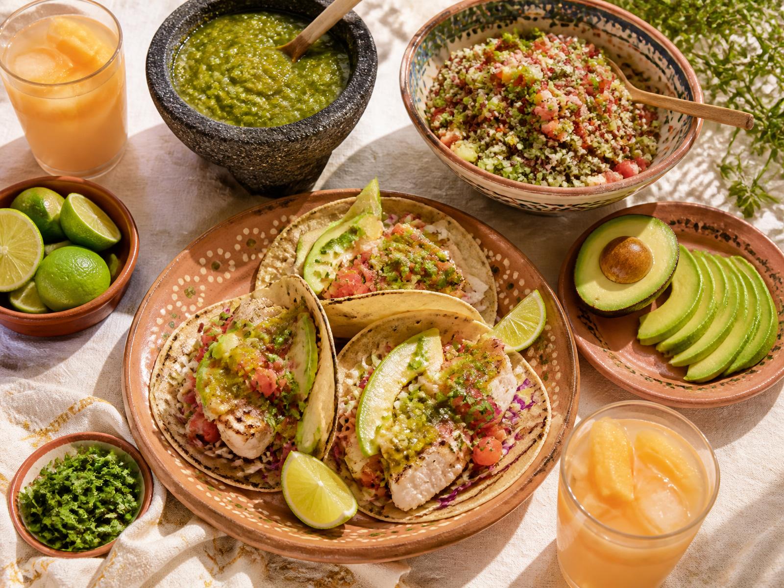 Mesa con tacos de pescado, salsa verde, aguacate y aguas frescas